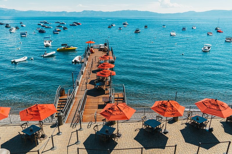 View of bright blue Lake Tahoe dotted with anchored boats with orange umbrellas over dining tables on a deck overlooking the lake