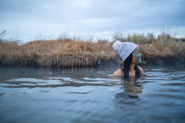 woman in hot spring with mud mask smiling wearing woolen cap